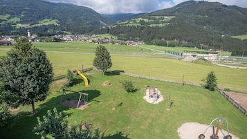 A playground with swings and a slide, surrounded by green fields and mountains in the background.