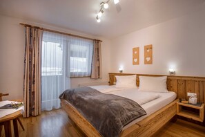 A bedroom with a wooden bed, a bedside table, a chair, and a window with sheer curtains.