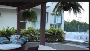 Un espacio de descanso al aire libre con sofás y cojines, rodeado de plantas en macetas y un edificio moderno al fondo.