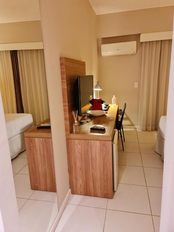 A hotel room with a wooden desk, a flat-screen TV, a lamp, and a minibar.