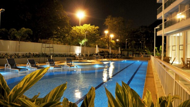 Una piscina nocturna con agua cristalina, rodeada de sillones de descanso y una valla blanca.