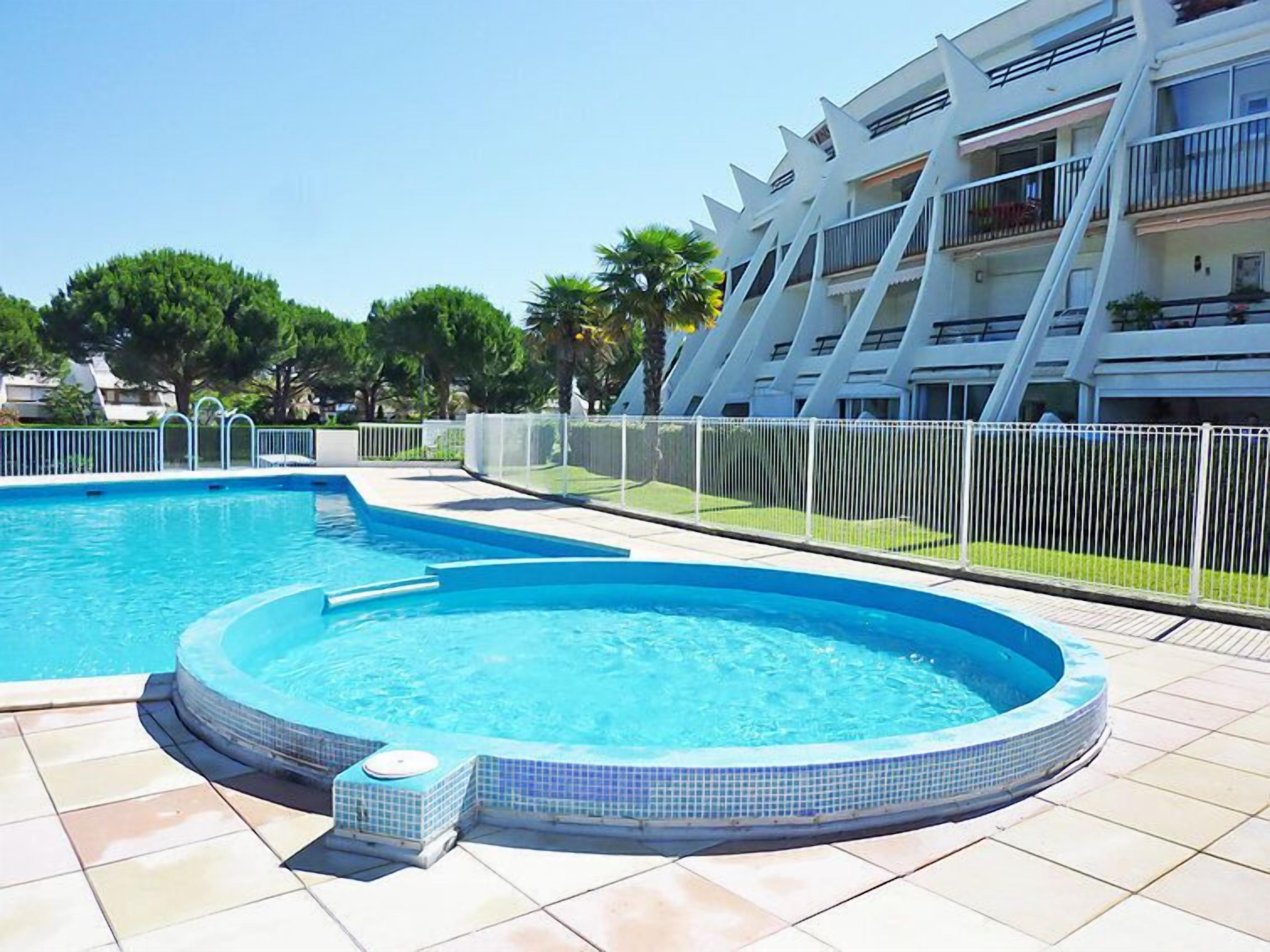 A swimming pool with a hot tub, surrounded by a white fence, and a modern building with balconies in the background.