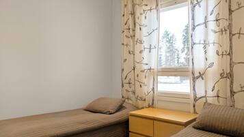 Una habitación pequeña y ordenada con dos camas individuales, una mesita de noche y una ventana con cortinas.