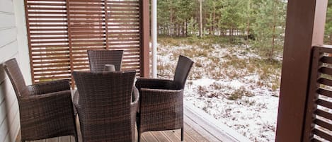 A covered outdoor area with a wooden deck, wicker chairs, and a view of a snowy landscape with trees.