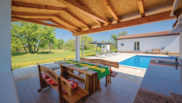 A covered outdoor area with a pool, wooden furniture, and a view of a house and trees.