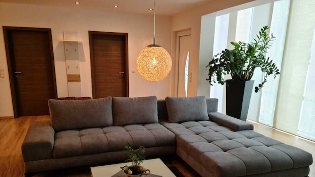 Een moderne woonkamer met een grijze modulaire bank, een witte salontafel met decoratieve objecten, een hanglamp en een grote potplant.