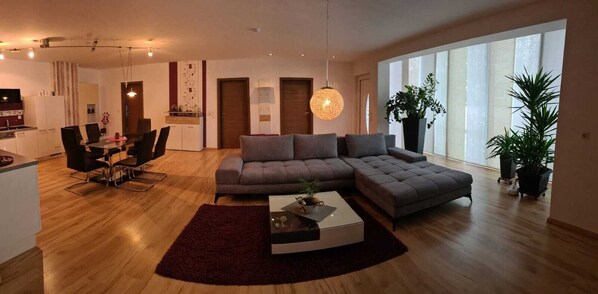 Un salon moderne comprenant un canapé d’angle, une table à manger avec des chaises, une table basse et de grandes fenêtres laissant entrer la lumière naturelle.