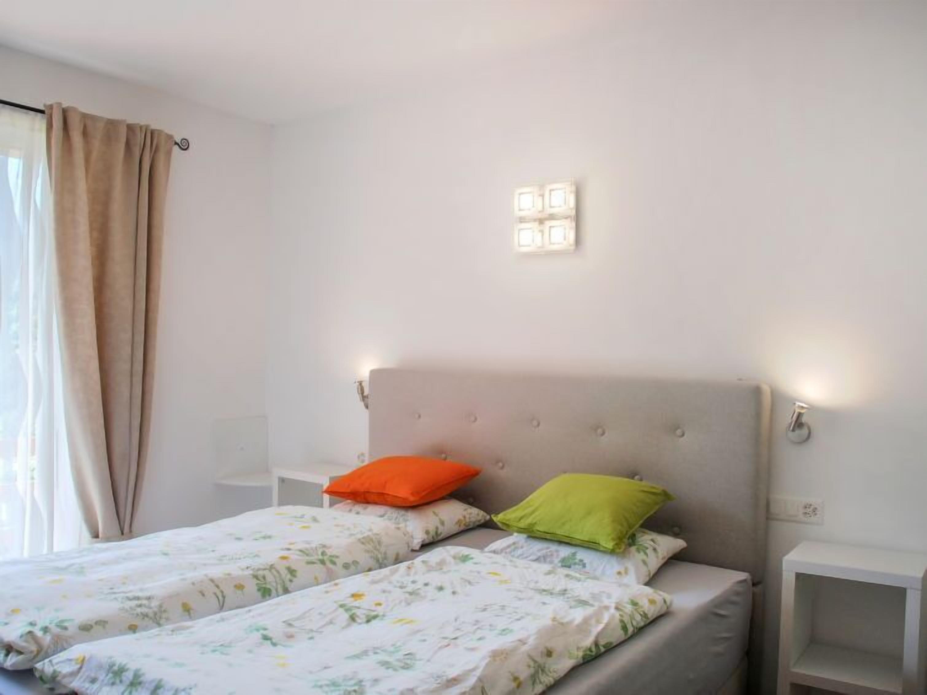 A double bed with floral bedding, two pillows, a headboard with light fixtures, and a window with curtains.