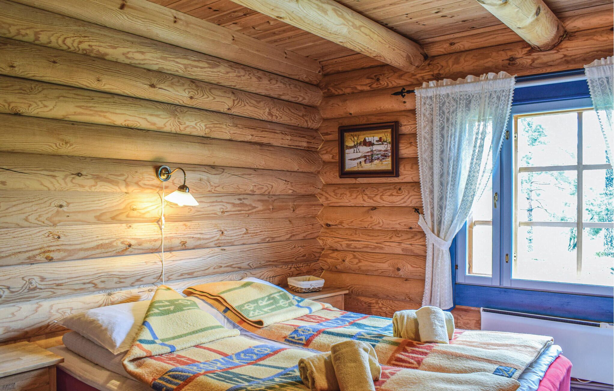 A wooden cabin room with a bed, a nightstand, a lamp, a window with curtains, and a painting on the wall.