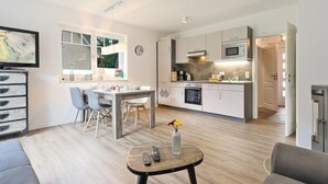 A modern kitchen with white cabinets, a microwave, and a stove. There is a dining area with a table and chairs, and a small round table with a vase and remote controls. - Reetdachhaus 20 auf Usedom (Zirchow/Usedom)