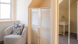 Una habitación con un radiador blanco, una ventana con persianas, un escritorio blanco con una lámpara y un armario blanco con puertas persianas.