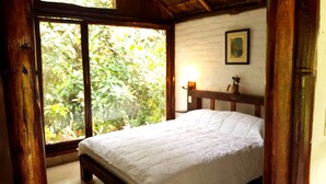 Un dormitorio con una cama de madera, un ventanal con vistas a la vegetación, una pared blanca y un cuadro enmarcado.