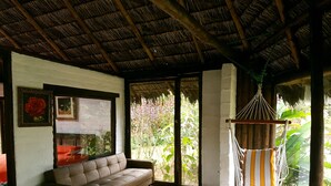 Una habitación con sofá, una hamaca, una ventana con vistas a la vegetación y un techo de paja.