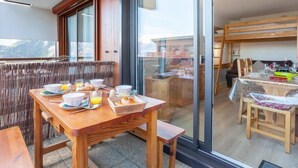 A dining area with a wooden table set for breakfast, a large window with a view, and a sliding glass door leading to a balcony. - Pegase Phenix by Interhome (Le Corbier)