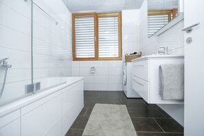 Un bagno moderno con box doccia in vetro, lavatrice integrata e un'ampia finestra con persiane in legno.