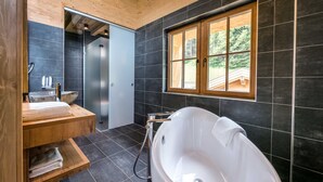 Une salle de bain moderne avec une baignoire sur pieds, des étagères en bois, une grande fenêtre et des murs recouverts de carreaux de couleur foncée.