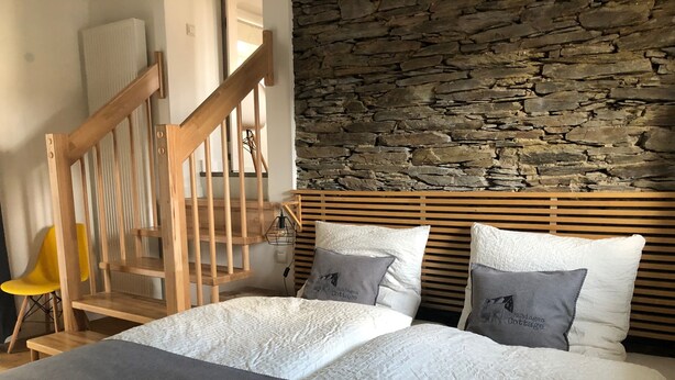 Quarto com parede de pedra, cabeceira de madeira e cama com roupa de cama branca e almofadas cinzentas.
