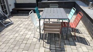 Uma área de pátio com mesa e cadeiras de metal, um banco e um pequeno canteiro de jardim.