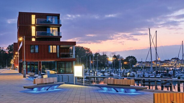 A modern multi-story building with balconies, a marina with boats, and a waterfront promenade with seating areas. - Passatvilla - Apartment 3 (Travemünde-Waterfront)