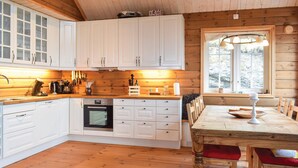 L’intérieur d’un chalet en bois est doté de meubles blancs, d’un coin cuisine avec des appareils électroménagers, d’une table à manger avec des chaises et d’une fenêtre donnant sur l’extérieur.