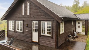 Une cabane en bois sombre, dotée d’un toit à pignon, d’une terrasse avec des sièges d’extérieur et d’un petit espace barbecue extérieur.