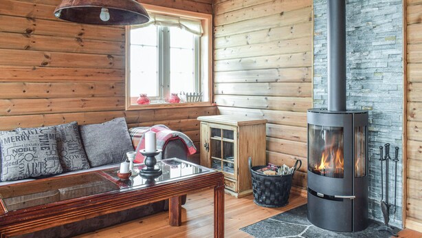 Un intérieur de chalet chaleureux, avec un revêtement mural en bois, une table basse en verre, un canapé avec des coussins, une cheminée en pierre et une fenêtre avec des rideaux.
