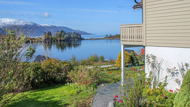 Et hus med en stenkø ved til en balkon med udsigt over en sø, hvor bjerge ses i baggrunden.