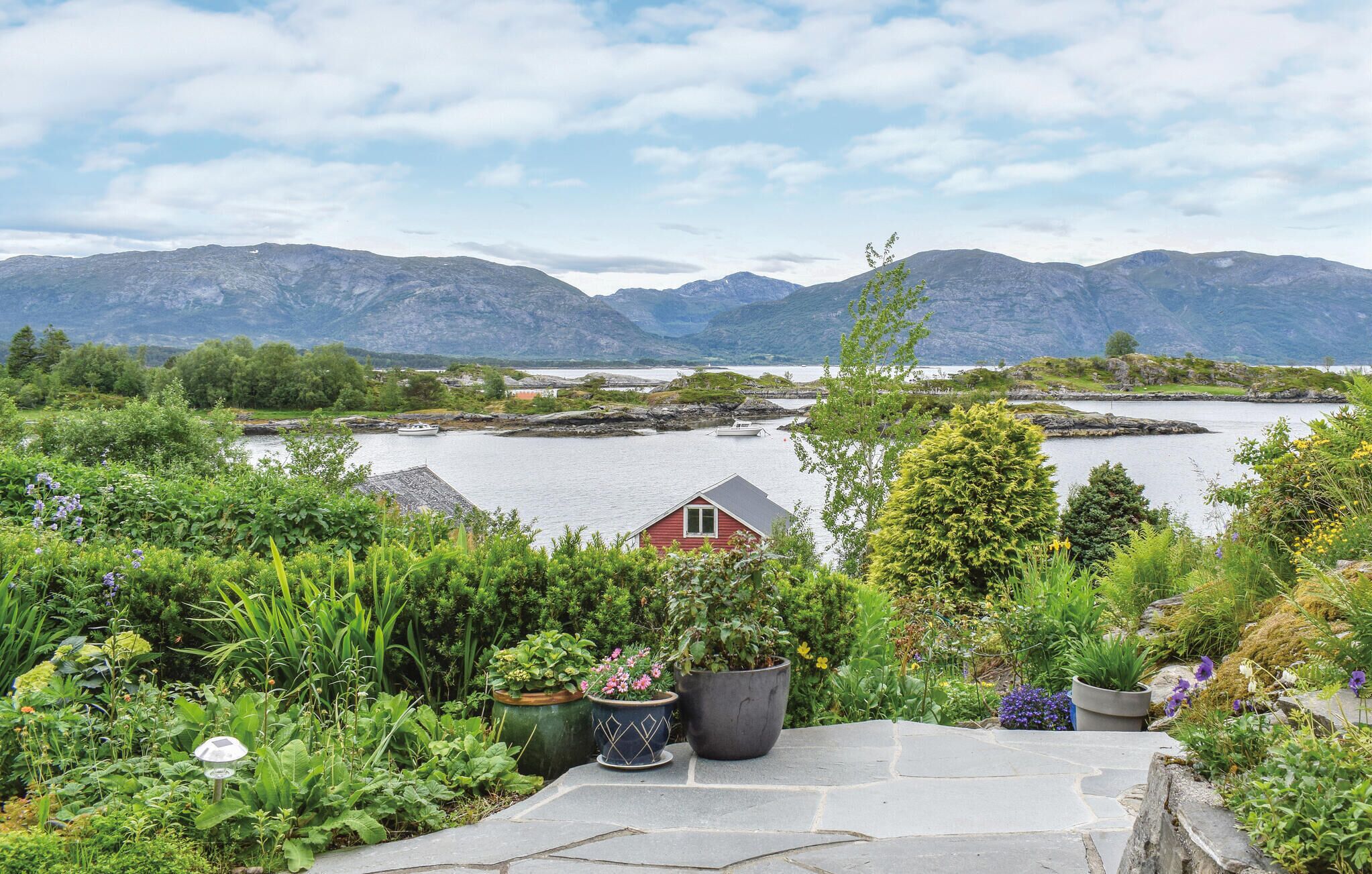 A scenic view of a lake with mountains in the background, a small red cabin, and a garden with various plants and flowers.