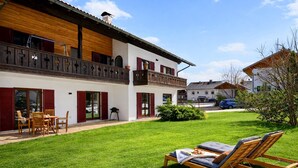 Une maison à deux étages avec un balcon, une pelouse dotée de chaises longues et une voiture garée dans l’allée.