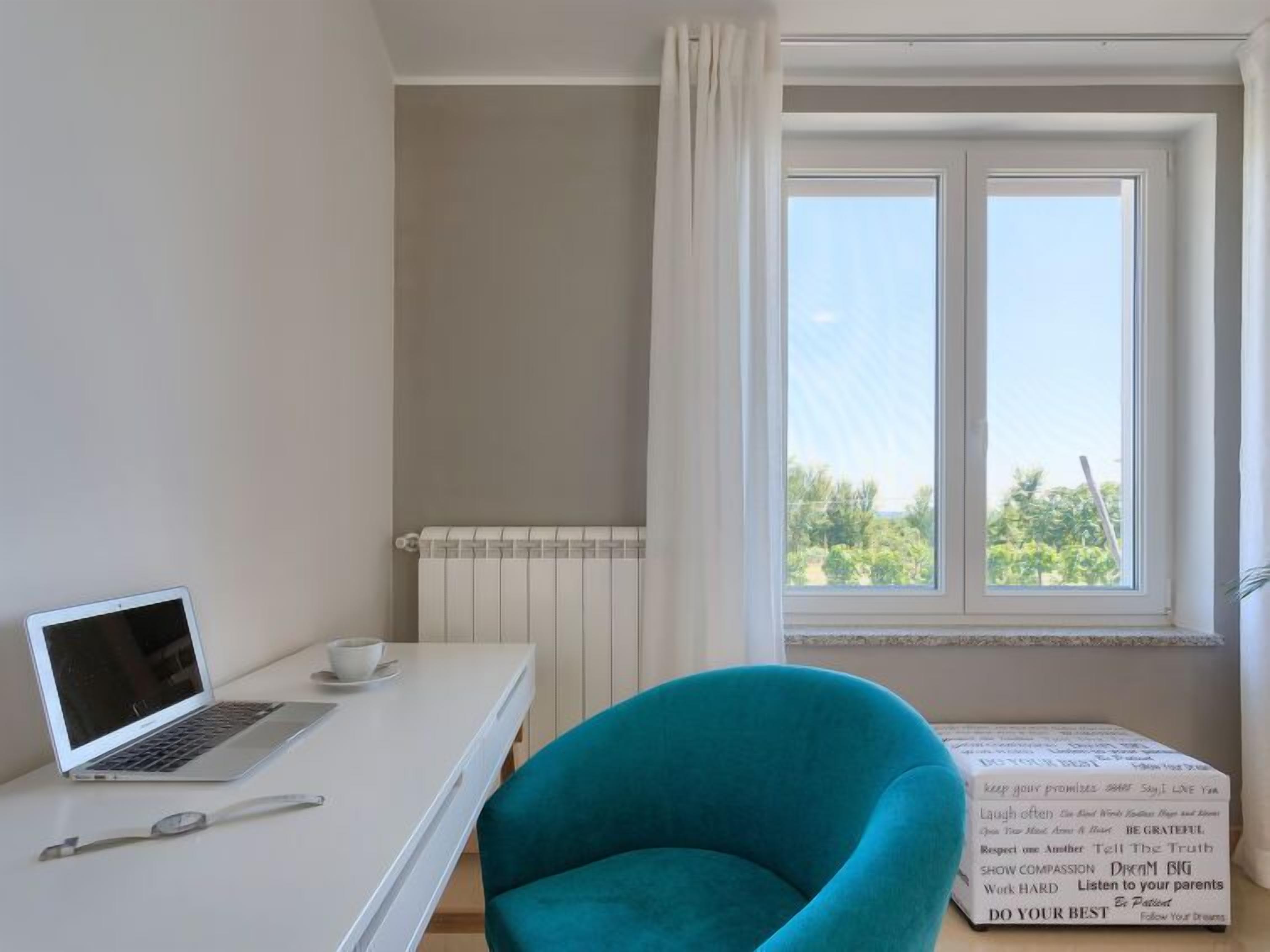 Une pièce avec un bureau blanc, un ordinateur portable, une tasse, une chaise et une fenêtre avec des rideaux.