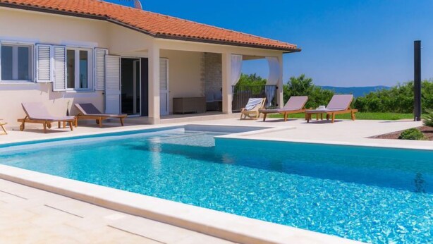 A poolside area with a clear blue pool, lounge chairs, and a building with a tiled roof. - Vacation home Eugen by Interhome (Barban)