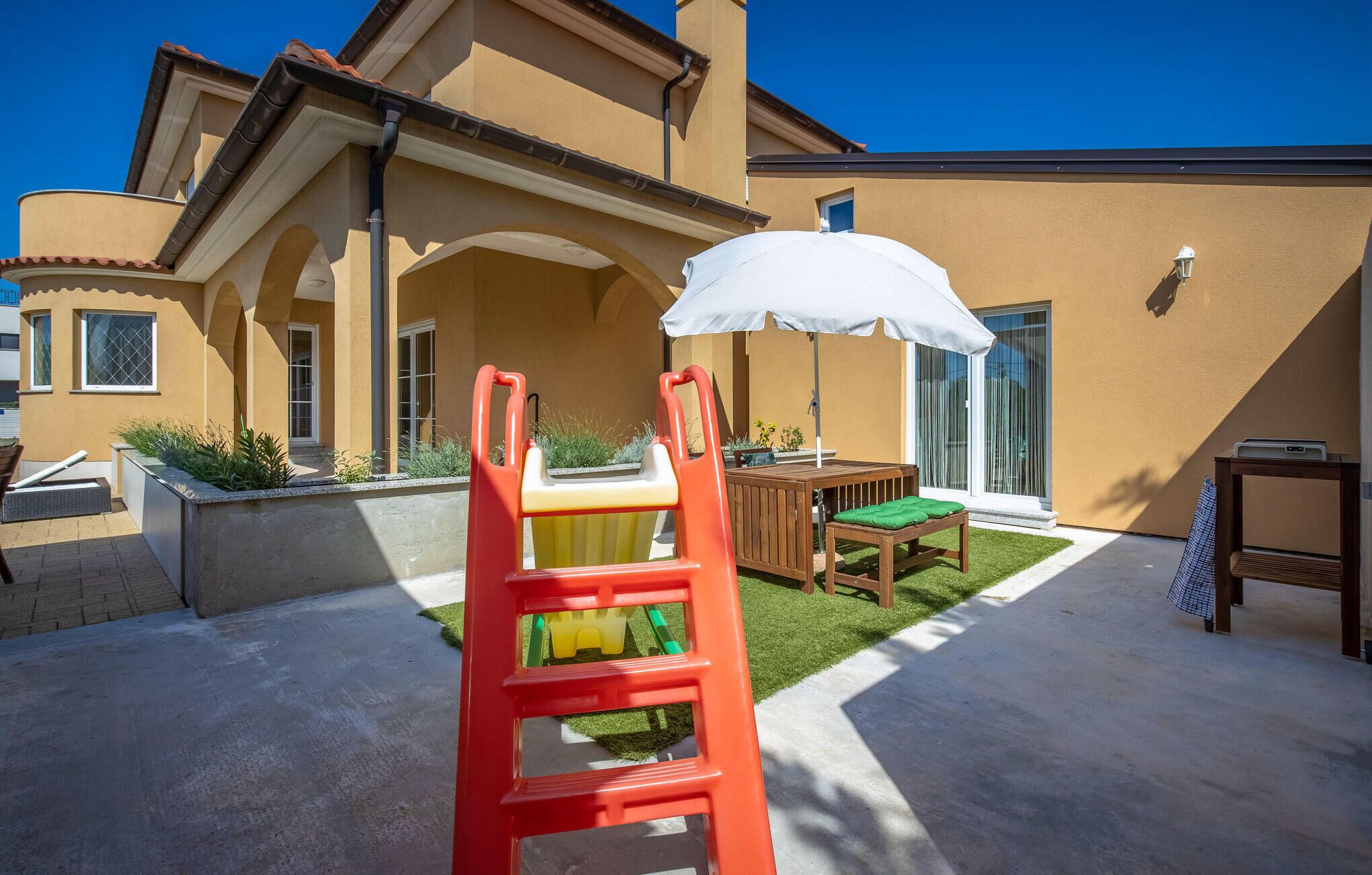 Un patio con una silla roja, una sombrilla blanca y una mesa de madera con un cojín verde.
