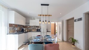 Una cocina moderna con armarios blancos, salpicadero de azulejos y un comedor con mesa de vidrio y sillas coloridas.
