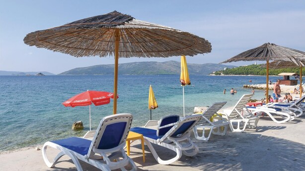 A beach with lounge chairs and umbrellas, clear blue water, and distant mountains. - Beautiful apartment in Seget Vranjica (Seget Vranjica)