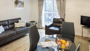 A modern living room with a leather sofa, a glass coffee table with a newspaper and fruit bowl, a leather armchair, a TV on a stand, and a large window with curtains. - Baggot Rath by Interhome (Dublin)