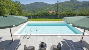 A rectangular swimming pool with a clear blue water surface, surrounded by lounge chairs and umbrellas, set against a backdrop of lush green hills and a clear sky. - Agr. Castello della Mugazzena by Interhome (Aulla)