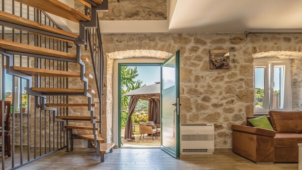Un'ampia stanza con una scala in legno, pareti in pietra, vista attraverso una porta aperta e un'area salotto con divano e poltrone.