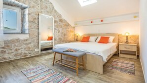 Una camera da letto con parete in pietra, un lucernario, un letto con biancheria bianca e cuscini arancioni, una panca, un comodino con una lampada e un tappeto colorato.