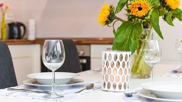 Een eettafel gedekt met borden, bestek en een glas water. Ook zichtbaar is een vaas met gele bloemen en een decoratieve kandelaar.