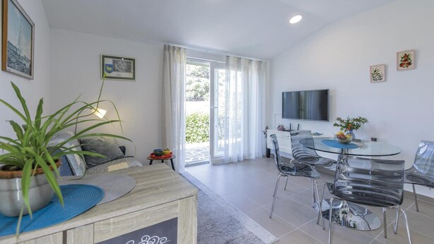 Een moderne woonkamer met een bank, een salontafel en een eethoek met een glazen tafel en stoelen. Er is een televisie aan de muur bevestigd en een deur die naar buiten leidt.