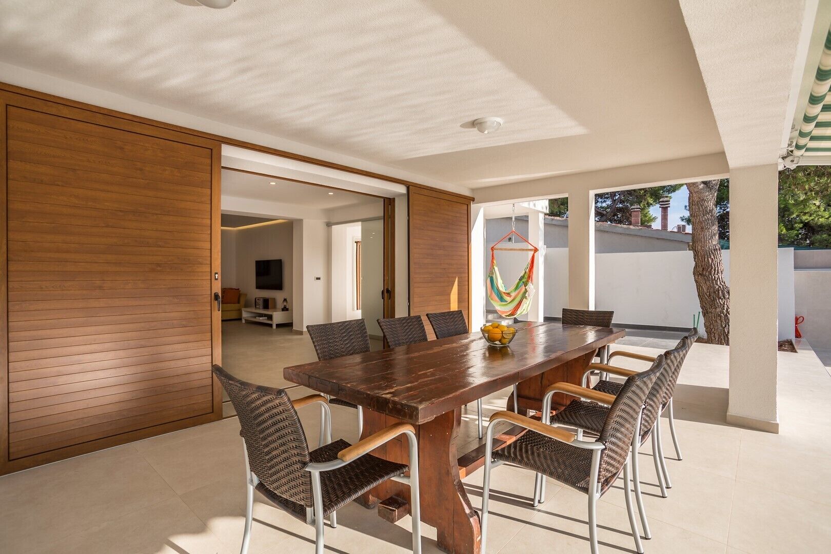 Un espace repas moderne avec une table en bois et des chaises en osier, un hamac suspendu et une vue sur l’extérieur.