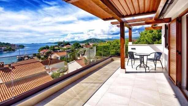 Un balcón con vista al mar y colinas, una pérgola de madera y una mesa con sillas.