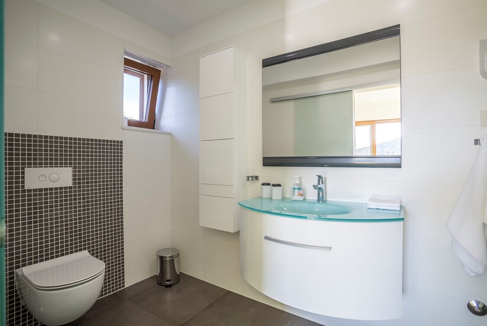 Ein modernes Badezimmer mit einem Glaswaschbecken, weißen Schränken, einer Toilette und einem Fenster.
