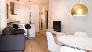 Un salon moderne avec un canapé gris, une table à manger blanche avec des chaises, un téléviseur posé sur un meuble blanc, et un coin cuisine équipé d’une cuisinière et d’un évier.