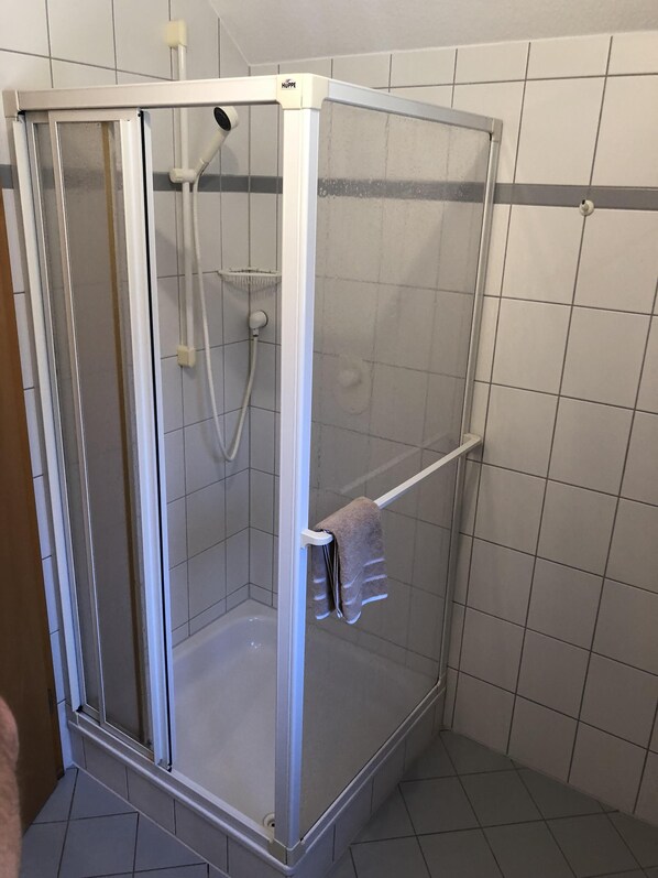 Box de chuveiro envidraçado com estrutura branca, chuveiro manual e porta-toalhas.
