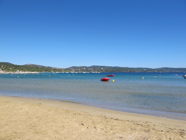 A beach with clear blue water, a sandy shore, and boats in the distance.
