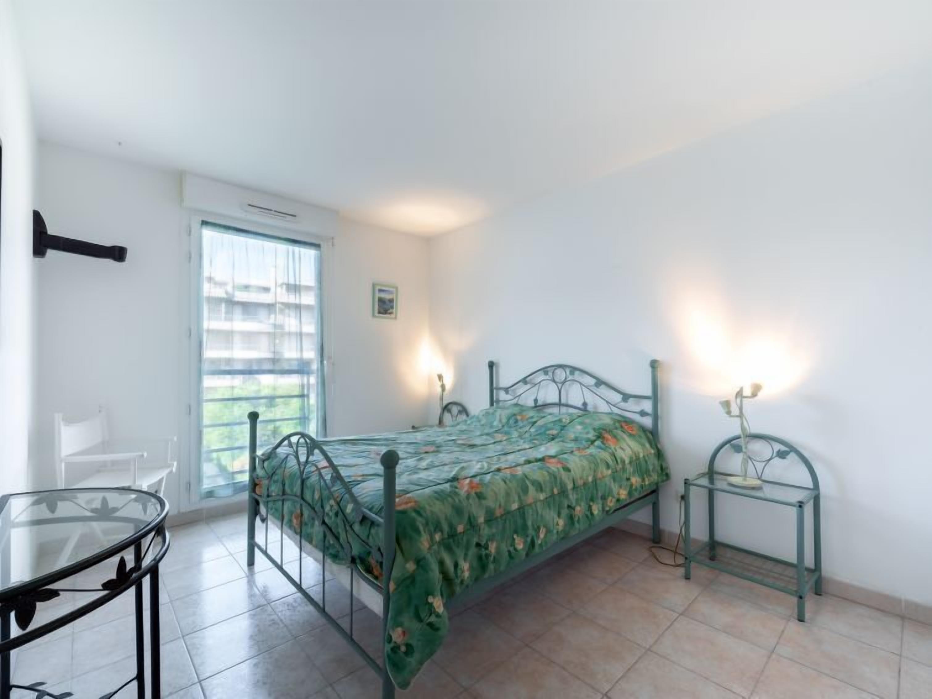 A bedroom with a bed, a glass table, a chair, a window with blinds, and a wall-mounted shelf.