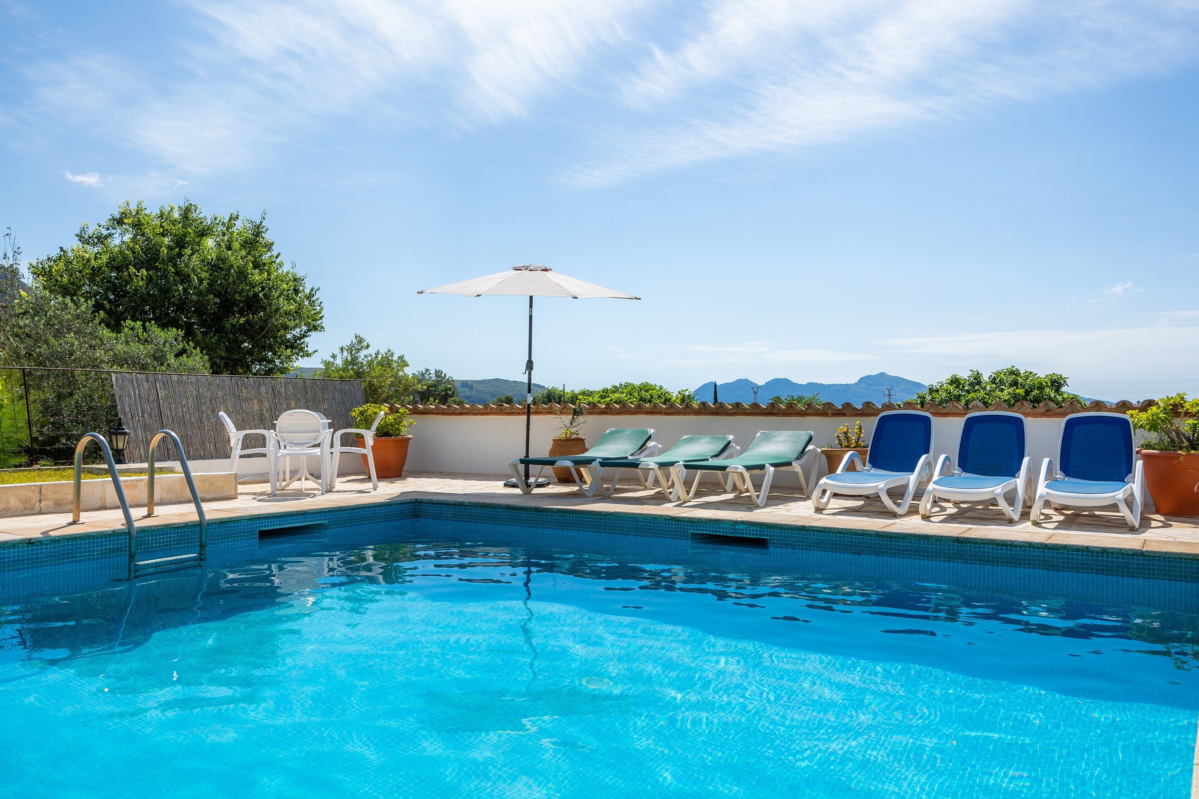 A swimming pool with lounge chairs and an umbrella, surrounded by greenery and mountains.