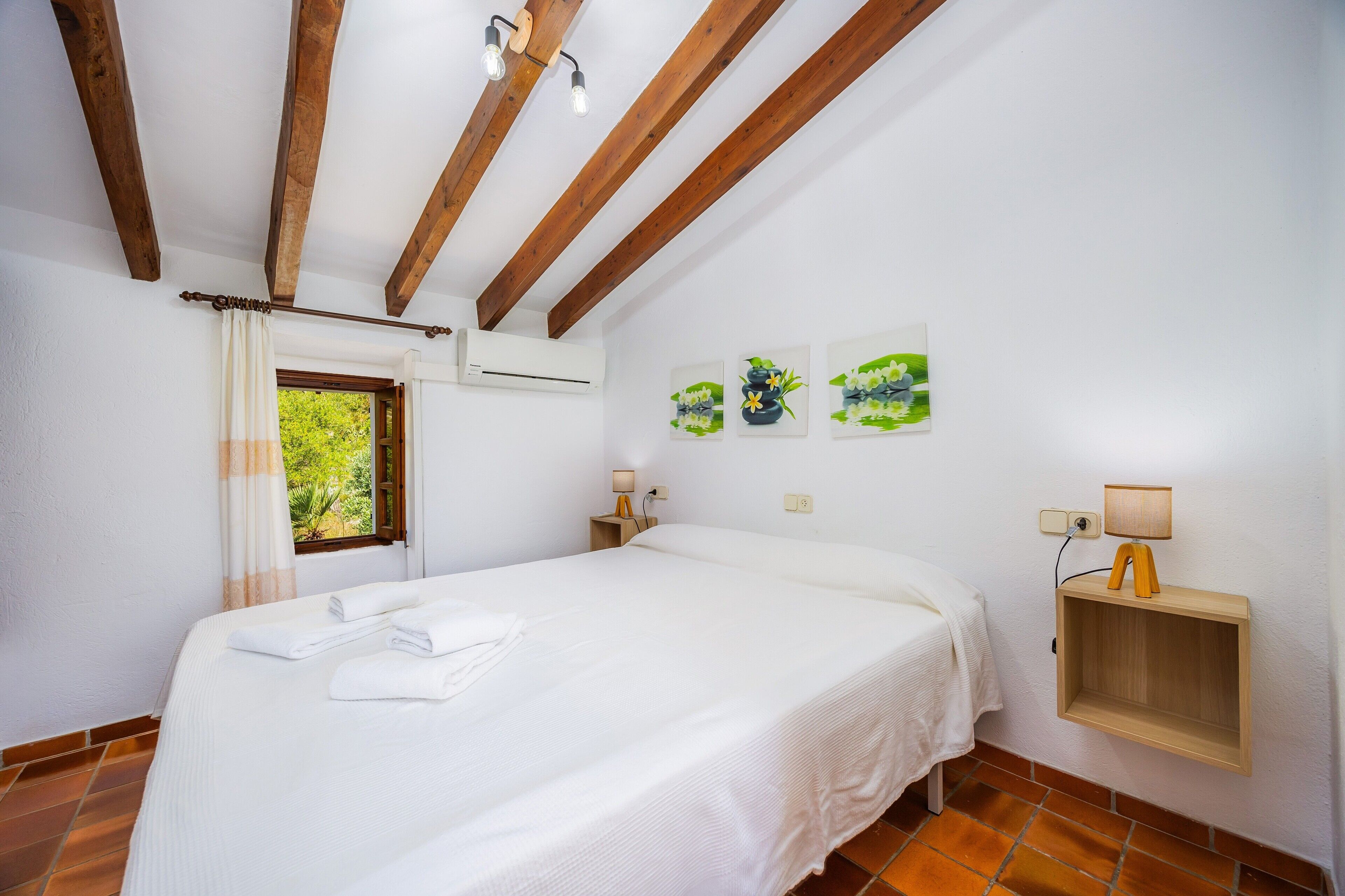 A bedroom with a bed, a window, a wall-mounted shelf with a lamp, and artwork on the wall.
