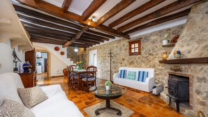 Un interior rústico con pared de piedra, vigas de madera, chimenea, una mesa redonda de vidrio y un sofá blanco.